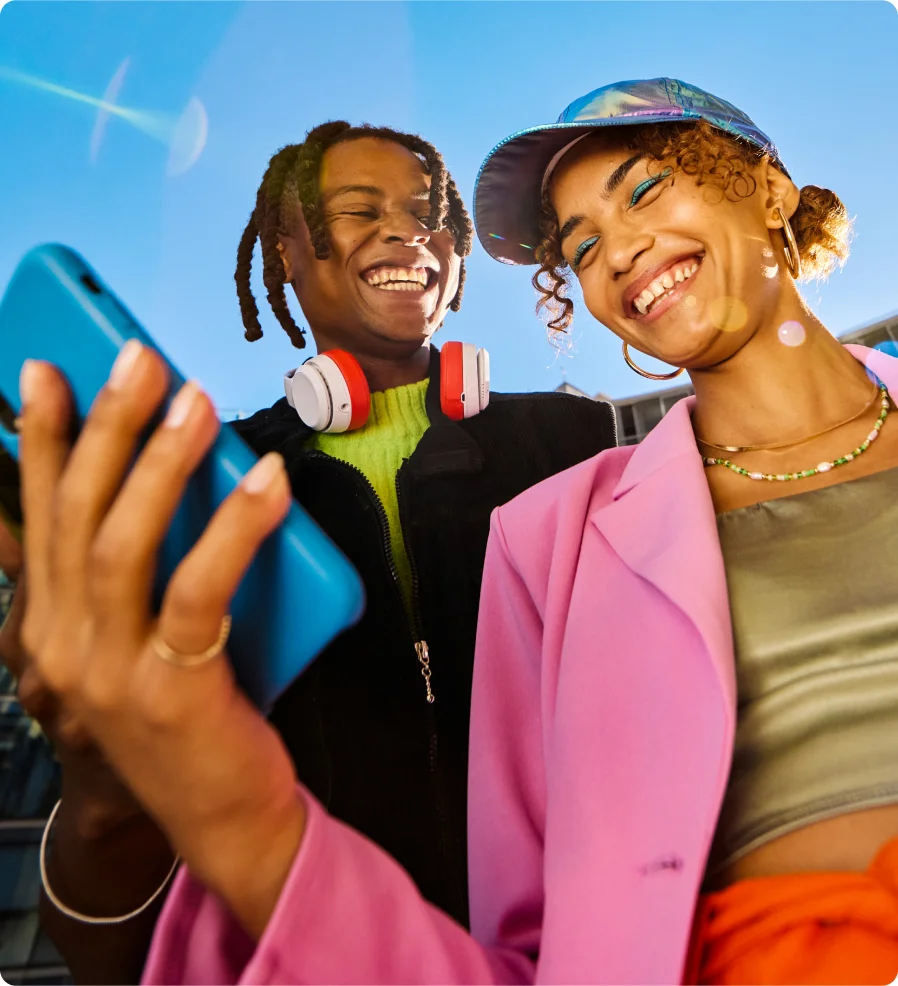 A woman smiling at her phone while holding it, with a man beside her also smiling at the screen.